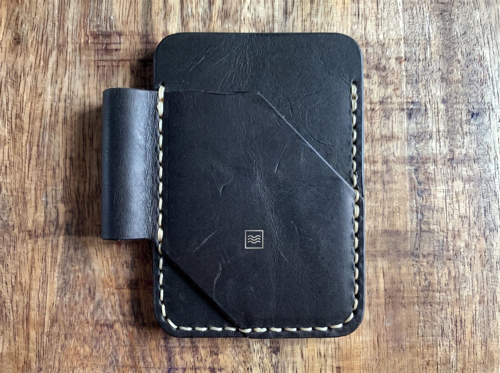 A card holder in black leather with an angled front pocket, a pen loop, cream saddle stitching and the Tidemark monogram in silver foil.